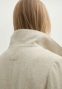 Bézs gyapjú keverék kabát magas gallérral, texturált felülettel és logóval a hátsó részen. Tiszta vonalak és testhezálló fazon.