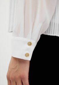 Weiße Bluse mit Ärmel aus zartem transparentem Stoff, plissiertem Design und doppelten goldenen Knopf-Akzenten an der Manschette. Keine sichtbaren Muster.