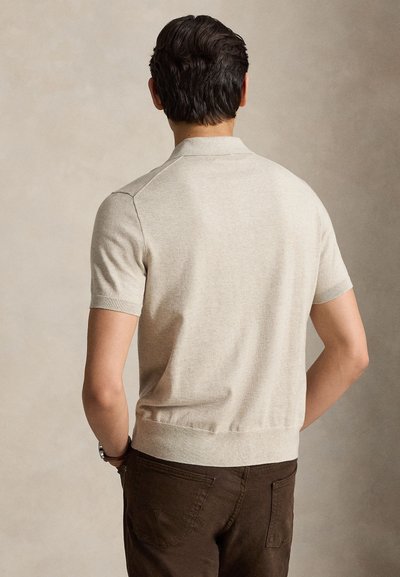 Άνδρας με σκούρα μαλλιά που φοράει μπεζ κοντομάνικο πουλόβερ και καφέ παντελόνια, στέκεται με την πλάτη στην κάμερα σε απλό φόντο.