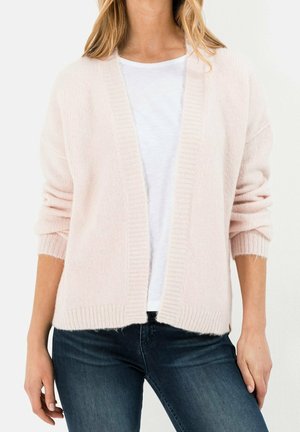 Frau trägt eine hellrosa offene Strickjacke über einem weißen T-Shirt und dunkelblauen Jeans vor einem schlichten hellen Hintergrund.