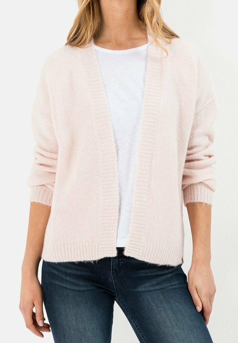 Femme portant un cardigan rose clair ouvert sur un t-shirt blanc et un jean bleu foncé, sur un fond clair uni.