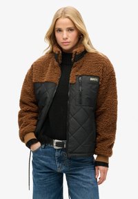 Veste en shearling marron avec un panneau inférieur noir matelassé, fermeture éclair à l'avant et une petite poche poitrine. Présente des poignets côtelés et une coupe moderne.