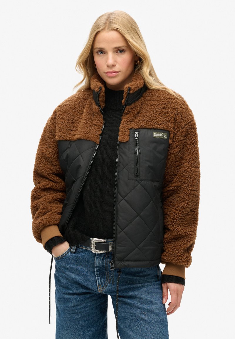 Veste en shearling marron avec un panneau inférieur noir matelassé, fermeture éclair à l'avant et une petite poche poitrine. Présente des poignets côtelés et une coupe moderne.