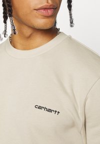 Sudadera beige de cuello redondo con un acabado acanalado, que presenta un pequeño logotipo negro bordado en el pecho. Material de algodón suave con un corte relajado.