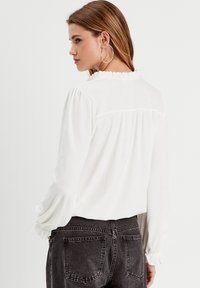 Weiße strukturierte Bluse mit Rüschenausschnitt und langen Ärmeln, kombiniert mit dunkelblauen Jeans. Hat geraffte Details an den Ärmelsäumen.
