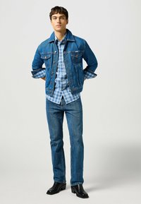 Denimjacka i medelblå, med två bröstfickor och knäppning med knappar, lagd över en ljusblå rutig skjorta. Kombinerad med mörkblå jeans och svarta stövlar.