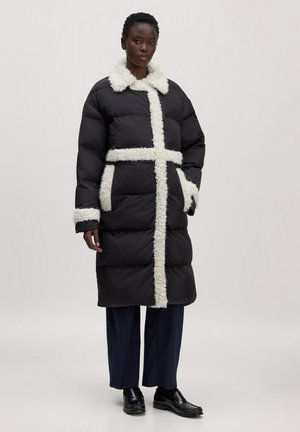 Long manteau matelassé noir avec bordure blanche duveteuse sur le col, le devant, les poignets et les poches, porté par-dessus un pantalon foncé et des chaussures noires.