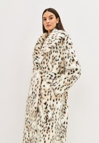 Hosszú sötét hajú nő fehér műszőrme kabátban, fekete és barna foltos mintával, amely kezét a zsebébe téve pózol egy sima háttér előtt.