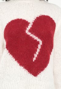 Adīts džemperis no mīkstas baltas dzijas ar lielu sarkana saplēsta sirds dizainu, kam ir teksturēts apdares elements un kontrastējošas adījuma detaļas.