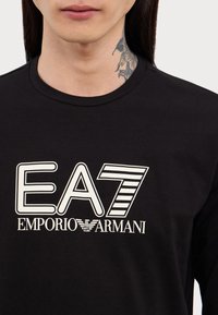 T-shirt in cotone nero con grande logo bianco "EA7 EMPORIO ARMANI" sul davanti, con una texture liscia e un design classico a girocollo.