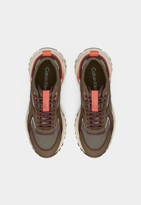 Ζευγάρι καφέ και μπεζ αθλητικά παπούτσια Calvin Klein με πορτοκαλί ταινίες έλξης και επενδεδυμένες ενδιάμεσες σόλες, σε θέα από ψηλά πάνω σε λευκό φόντο.