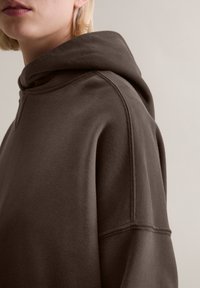 Kapuzensweatshirt in Dunkelbraun aus strukturiertem Stoff. Mit lockerem Schnitt, überschnittenen Schultern und einer markanten Kapuze.