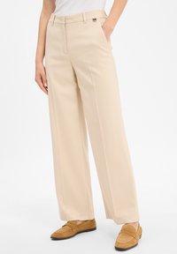 Woman wearing beige wide-leg trousers with side pockets and light brown suede loafers, standing against a white background.