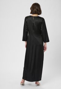 Frau mit kurzem braunem Haar, die ein langes schwarzes Satin-Kleid und hellbeige Absätze trägt und mit dem Rücken zu einer neutralen Hintergrund steht.