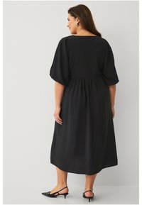 Zwarte midi-jurk met korte, uitzwenkende mouwen, een taille die getailleerd is en een vloeiende silhouet. Draagt zwarte hakken, waardoor een gestructureerde stof wordt getoond.