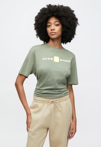 Πράσινο βαμβακερό μπλουζάκι με κοντά μανίκια, με κίτρινο και λευκό λογότυπο "NIKE SHOX", συνοδευόμενο από μπεζ φόρμα με κορδόνι στη μέση.