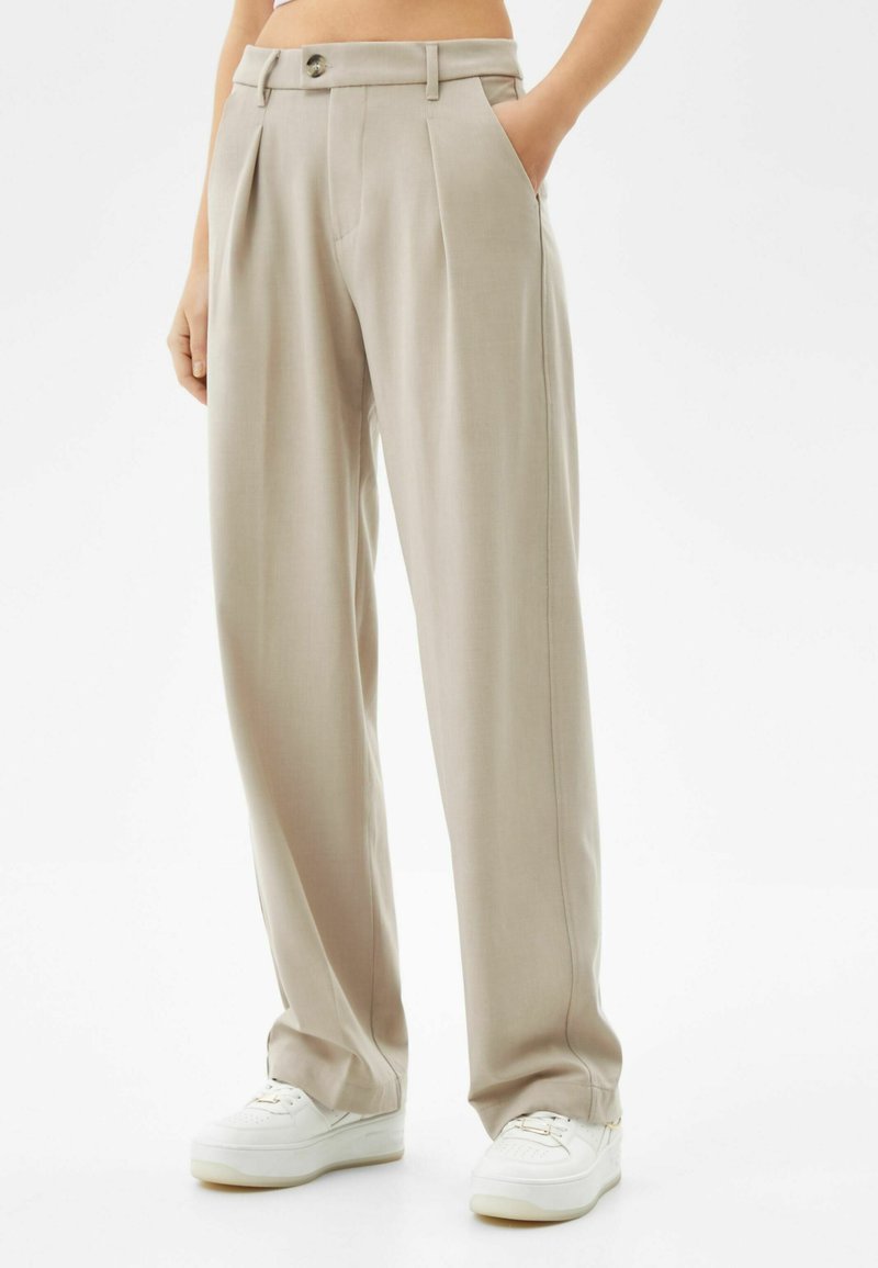 Beige, geplooide broek met een hoge tailleband, voorzien van zijzakken en een enkele knoopsluiting; gedragen met witte sneakers.