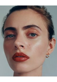 Lähikuva kalpeaihoisesta kasvoista, sinisistä silmistä, täyteläisistä huulista tummanpunaisessa kiillossa, kaarevista kulmakarvoista ja pienestä kultaisesta renkorengaskorvakorusta. Sileä pinta.