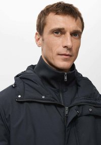 Giacca nera imbottita con colletto alto, caratterizzata da una chiusura con zip, bottoni a pressione e una finitura testurizzata. Cappuccio regolabile e tasche laterali.