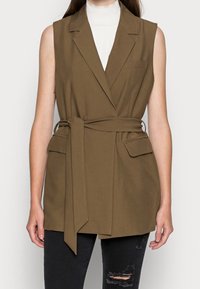 Brun ärmlös blazer med bälte i midjan, snedskurna slag och framfickor, bärs över en vit höghalsad topp. Texturerad tyg.