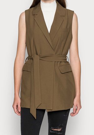 Blazer senza maniche marrone con vita cintata, rever a punta e tasche frontali, indossato sopra un top bianco a collo alto. Tessuto testurizzato.
