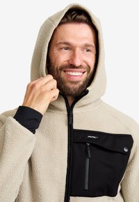 Beige fleece hoodie med en svart dragkedjeficka, dragkedjestängning och justerbar huva. Har strukturerad tyg och kontrasterande svarta detaljer.