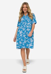 Vestido azul de manga corta con patrones blancos similares a corales, con escote en V y corte holgado. Combinado con sandalias blancas de tacón cuadrado.