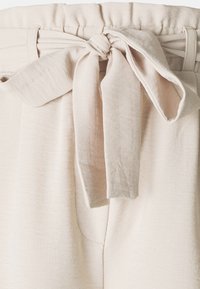 Gros plan sur un pantalon en tissu beige avec une ceinture élastique et un nœud décoratif noué à l'avant au centre.