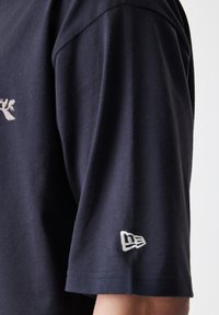 Temno modra majica s kratkimi rokavi iz bombaža, z belim grafičnim motivom na prsih in vezenim logotipom na levem rokavu.