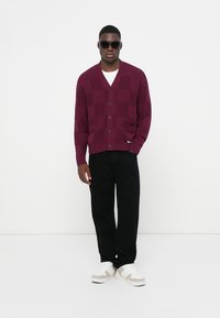 Cardigan a maglia bordeaux con motivo a scacchi, bottoni e tasche, abbinato a pantaloni neri e sneakers bianche. Uomo che indossa occhiali da sole.