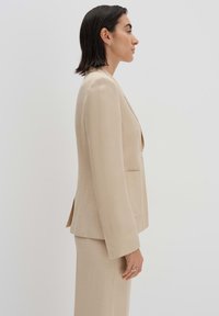 Blazer beige su misura realizzato in tessuto leggero, dotato di chiusura a un bottone e due tasche frontali, con una silhouette strutturata.