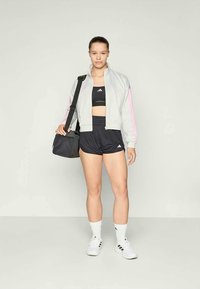 Ung kvinne i treningstøy, inkludert sports-bh, shorts, jakke og joggesko, som holder en svart treningsbag over skulderen.