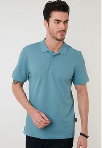 Polo de culoare teal cu guler clasic, mâneci scurte, placket cu trei butoane și un detaliu mic pe lateral, fabricat dintr-un material neted din amestec de bumbac.