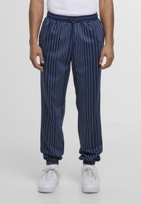 Pantaloni della tuta blu navy con strisce bianche verticali, vita elastica con cordino e polsini affusolati alle caviglie. Indossati sneaker bianche.