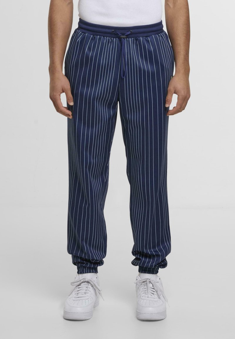 Pantaloni della tuta blu navy con strisce bianche verticali, vita elastica con cordino e polsini affusolati alle caviglie. Indossati sneaker bianche.