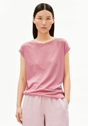 Frau mit glatten dunklen Haaren, die ein rosa T-Shirt mit hochgekrempelten Ärmeln und hellrosa Hosen trägt, steht mit einer Hand in der Tasche vor weißem Hintergrund.