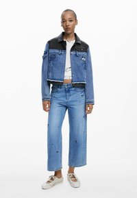 Giacca di jeans tagliata con dettagli in pelle nera, jeans azzurri chiari con ricami, e sneaker bianche con finitura strutturata.
