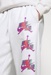 Bílé tepláky s třemi velkými potisky loga "Nike Air" v purpurové a zelené barvě, vyrobené z měkkého materiálu s nastavitelným pasem na tkaničku.