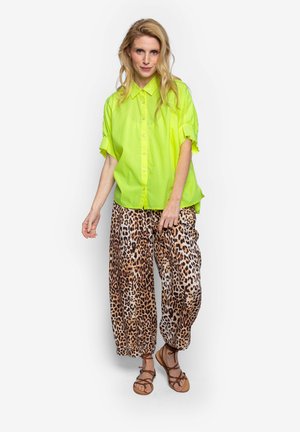 Blonde vrouw die een felgele korte mouwen overhemd met knopen draagt, wijde broek met luipaardprint en bruine sandalen met bandjes, staand tegen een witte achtergrond.