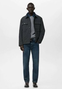 Donkergrijze jas met twee borstzakken en een blauwe kraag, gedragen over een grijze trui en blauwe jeans; zwarte laarzen maken de outfit compleet.