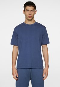 Tummansininen puuvillapaita lyhyillä hihoilla, pyöreällä kaula-aukolla ja hienovaraisella brändäyksellä vaaleansinisessä rinnassa. Sujuva rakenne, rento istuvuus.