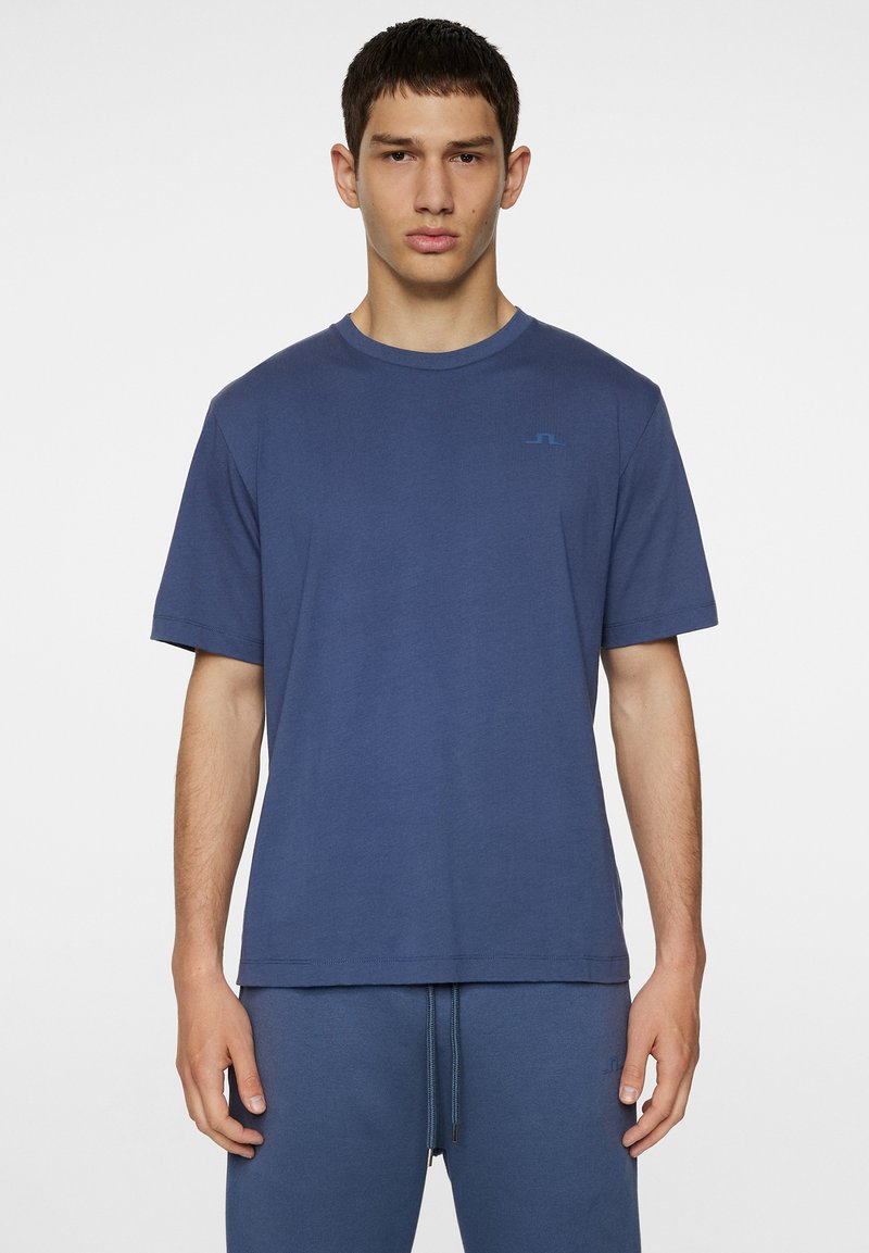 Tummansininen puuvillapaita lyhyillä hihoilla, pyöreällä kaula-aukolla ja hienovaraisella brändäyksellä vaaleansinisessä rinnassa. Sujuva rakenne, rento istuvuus.