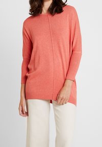 Koronkowy, oversize sweter w kolorze koralowym, z luźnym krojem, ryflowanymi mankietami oraz detalem wzdłuż środkowego szwu. Miękka faktura, idealna na codzienne wyjścia.