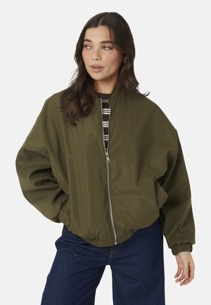 Vrouw draagt een olijfgroene jas met rits, elastische boorden en zoom, donkerblauwe spijkerbroek en een gestreept hemd eronder, kijkt naar links.
