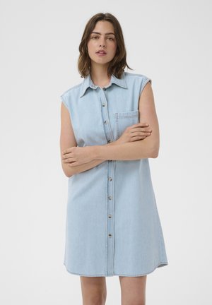 Kobieta o brązowych włosach do ramion, ubrana w bezrękawową, jasnoniebieską sukienkę dżinsową na guziki z kieszenią na piersi, stojąca z założonymi ramionami.