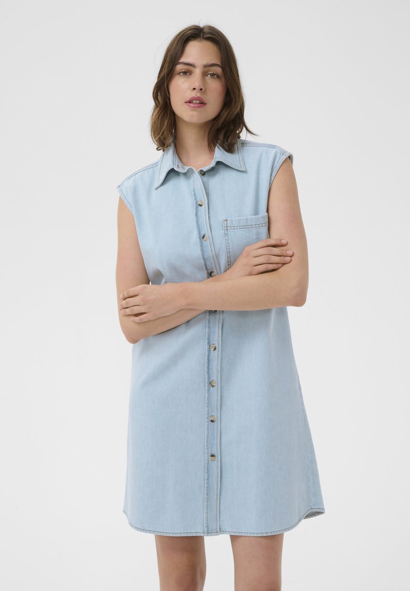 Femme aux cheveux bruns mi-longs portant une robe en jean bleu clair sans manches, boutonnée avec une poche poitrine, debout les bras croisés.