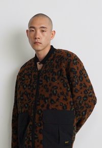 Jakke i brun fleece med leopardprint, sort lynlås, to forlommer og sort krave. Blød tekstur og oversized design.