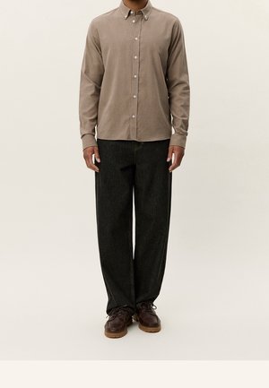 Mężczyzna w beżowej, zapinanej na guziki koszuli z długim rękawem, luźnych czarnych spodniach i brązowych butach sznurowanych, stojący na tle jednolitego tła.