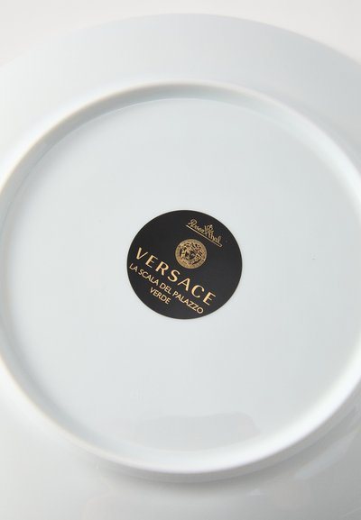 Λευκό πορσελάνινο πιάτο με λεία επιφάνεια. Διαθέτει μαύρη κυκλική ετικέτα με χρυσά γράμματα που αναγράφουν "Versace" και το όνομα του σχεδίου.