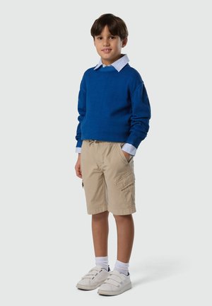 Niño de pie con un suéter azul, camisa a rayas, pantalones cortos cargo beige, calcetines blancos y zapatillas blancas frente a un fondo liso.